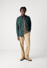 Camisa verde de botones sobre un suéter marrón, pantalones beige y zapatos color crema. Tela suave, ajuste entallado, con botones visibles y un logo sutil.