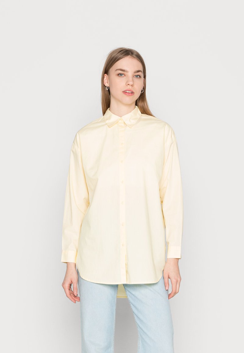 Camisa de botão feita de tecido leve na cor amarela suave, apresentando um colarinho clássico, mangas longas e uma bainha reta.