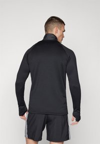 Haut de sport noir à manches longues avec col haut, texture lisse et design ajusté, associé à un short noir assorti avec des rayures blanches sur les côtés.