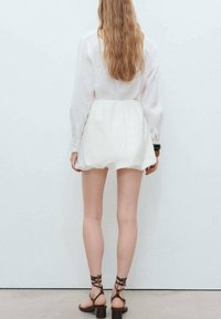 Vestito bianco corto con maniche lunghe e vita cintrata, realizzato in tessuto leggero. Abbinato a sandali marroni con cinturini, vista da dietro.