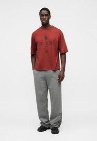 T-shirt en coton rouge avec un motif "ULTRAS" sur le devant, associé à un pantalon de survêtement gris avec un détail logo, porté avec des baskets noires.