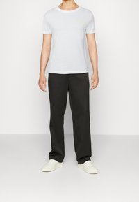 T-shirt bianca in cotone con collo tondo e scritte gialle, abbinata a pantaloni scuri e sneakers bianche, indossata da un modello.