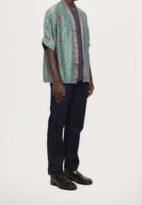 Mønstrede turkise kimono-jakke med korte ærmer, kontrastfarvet mønstret kant, båret over en mørk skjorte, parret med mørke jeans og sko.