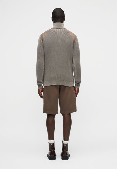Άνδρας που φοράει πουλόβερ πλεκτό ταυτίν χρώματος με σουέντ επιθέματα στους ώμους, καφέ σορτς μήκους μέχρι τα γόνατα, λευκές κάλτσες και σκούρα καφέ μπότες, στέκεται με την πλάτη στραμμένη.