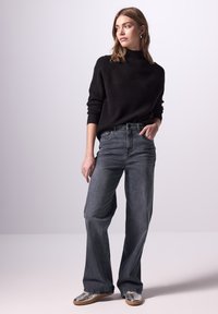 Zwarte geribbelde trui, hoge hals, gecombineerd met hoge taille donkergrijze brede jeans. Het model draagt metalic schoenen met een subtiele print.