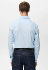 Camisa de rayas azul claro de manga larga con detalle en el yugo trasero, metida dentro de unos jeans de mezclilla oscuros, con un cinturón negro. Textura suave y diseño clásico.