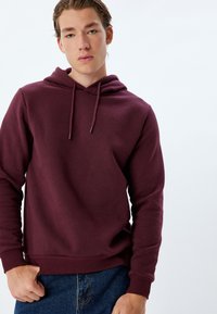 Felpa bordeaux realizzata in tessuto morbido con tasca a marsupio, cordini regolabili e polsini e orlo a costine. Abbinata a jeans blu.