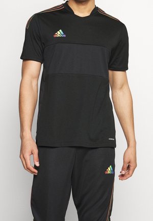Man draagt zwarte Adidas sportkleding met regenbooggestreepte logo's op de borst en dij, en regenboogstrepen op de schouders en zijkanten van de broek.