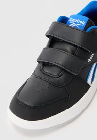 Sapatilha de couro preta com detalhes em azul, apresentando duas tiras de Velcro, detalhe perfurado na biqueira e uma sola de borracha branca.