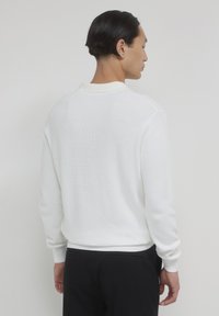 Mann, der mit dem Rücken zur Kamera steht, trägt einen weißen Strickpullover und schwarze Hosen vor einem schlichten hellen Hintergrund.