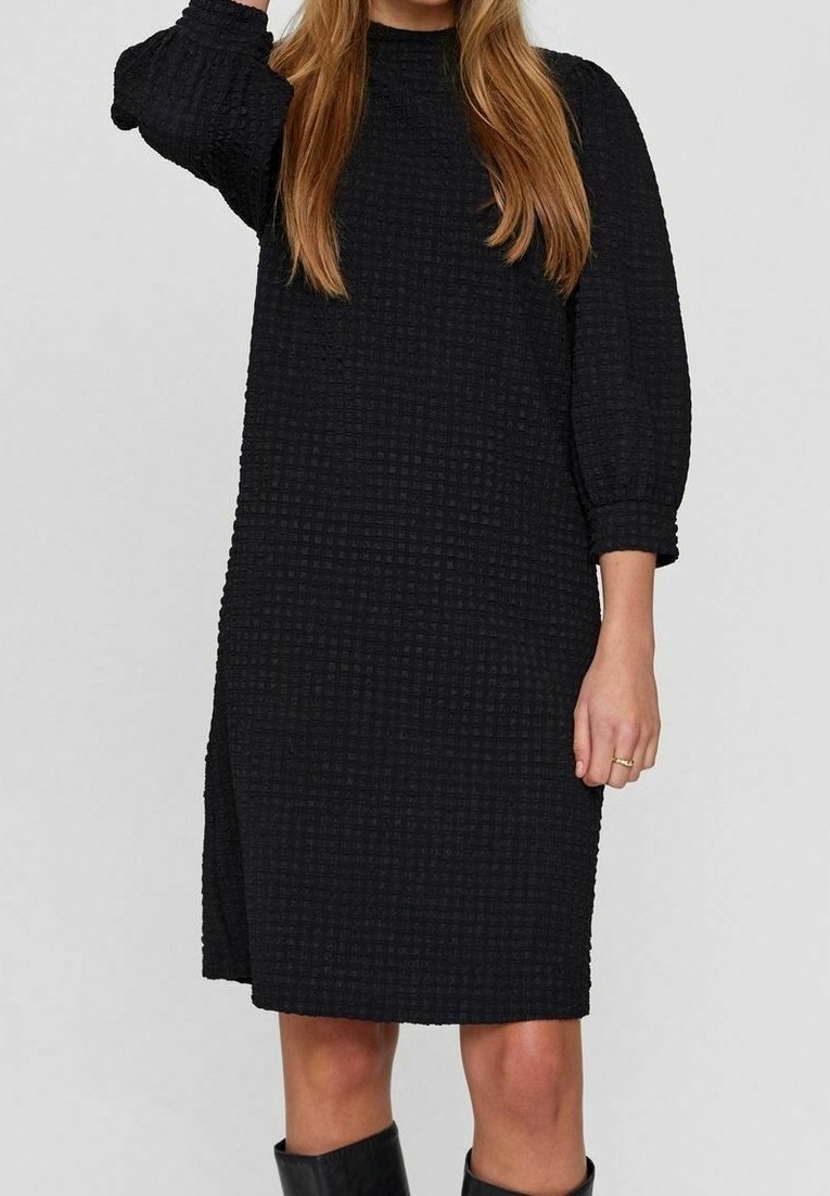 Svart texturerad klänning med hög halsringning, lösa trekvartsärmar och sidoklyfta, tillverkad av ett vävt tyg i rutmönster.