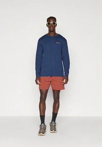 Tamnoplava majica dugih rukava s logotipom, kratke hlače boje hrđe, crne čarape do gležnja i smeđe višematerijalne cipele s vezicama i uzorkovanim potplatima.