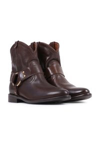 Een paar donkerbruine leren enkellaarsjes met een band en gespsluiting, een lage geprofileerde hak en afgeronde neuzen op een witte achtergrond.