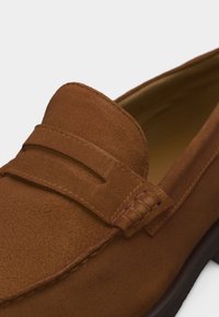Gros plan sur un mocassin en daim brun avec un détail de bande cousue et une doublure intérieure en cuir lisse.