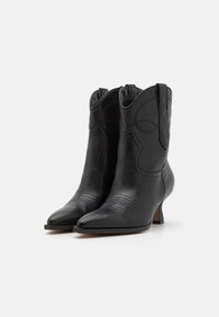 Botas de vaquero de cuero negro con punteras afiladas, detalles de costura en la parte delantera, tacones bajos y acentos decorativos en la parte superior del caña.