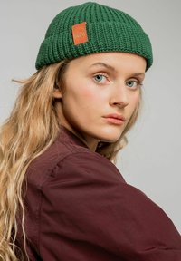 Zielona dziergana czapka typu beanie z teksturowanym wzorem i brązową skórzaną łatą logo, noszona z ciemnoczerwonymi koszulą, z długimi falującymi włosami.