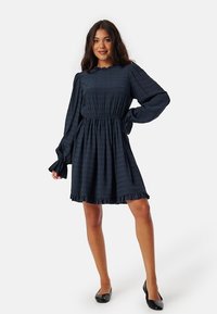 Robe bleu marine en tissu texturé, manches longues bouffantes et ourlet volanté. Taille élastique pour une silhouette ajustée. Des ballerines complètent le look.