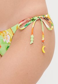Parte inferior de bikini floral em verde com detalhes em laranja e amarelo. Apresenta cordões laterais com contas decorativas e um pequeno charme de banana amarela.