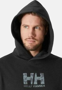 Sudadera negra con un logo de Helly Hansen en camuflaje en la parte delantera, que presenta una gran capucha y una tela suave y texturizada.