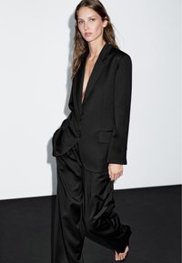 Kvinde iført en løs sort blazer uden skjorte under og matchende vidde-bukser, stående barfodet mod en lys væg.