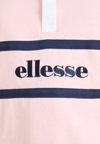 Polo majica od svijetloružičastog materijala s tamnoplavim prugama. Bijela ovratnik i izvezeni "ellesse" logo u tamnoplavoj boji. Mekana tekstura, ležeran dizajn.