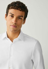 Biela košeľa s dlhým rukávom, klasickým golierom, zapínaním na gombíky a hladkou textúrou, ktorú nosí osoba s krátkymi kučeravými vlasmi na neutrálom pozadí.