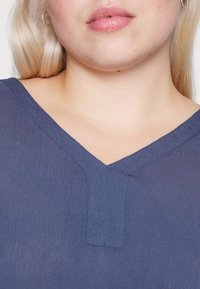 Námořnická modrá V-krk blůza s texturovanou tkaninou a malou detailní tabulkou u výstřihu. Rty v barvě pleti a světlé vlasy jsou částečně viditelné.