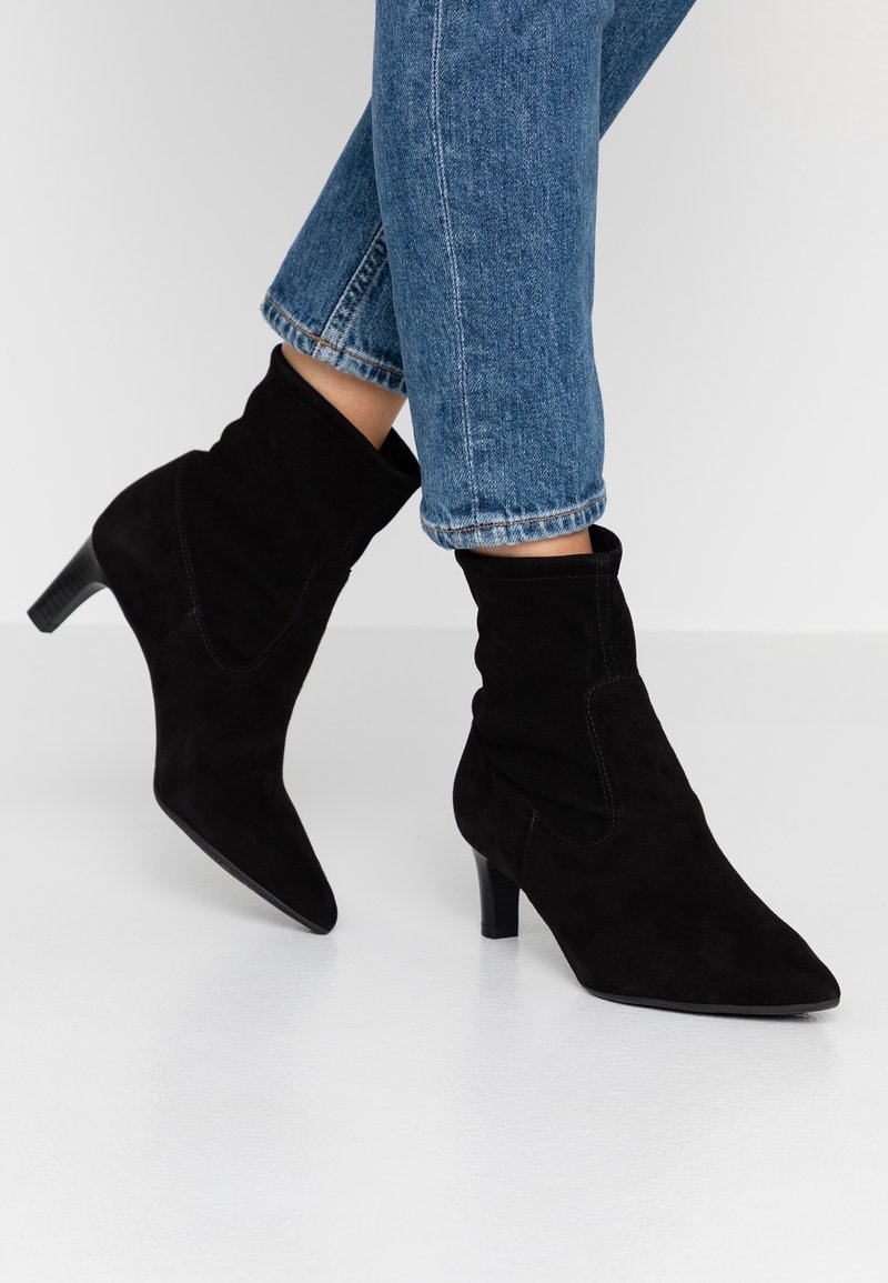 Personne portant des bottines en suède noir à talons moyens et un jean en denim bleu, debout sur un sol blanc lisse.