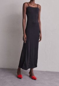 Czarna, bezrękawnikowa sukienka maxi o kwadratowym dekolcie i gładkiej fakturze, w połączeniu z czerwonymi, obcasami mules z frędzlami.