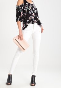 Top off-the-shoulder nero a fiori, jeans skinny bianchi, tacchi aperti neri, che tiene in mano una borsa a tracolla rosa pallido con accessori in argento.