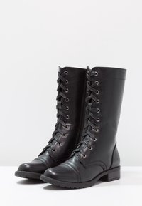 Botas de cuero negras hasta la rodilla con punta redonda, cordones en la parte delantera y cremallera lateral. Suela gruesa y tacón bajo apilado. Textura suave con detalles de costura.