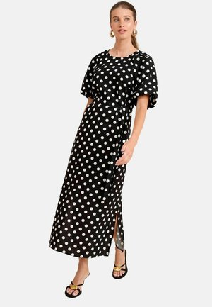 Vestido maxi negro con lunares blancos, mangas cortas abullonadas, cinturilla y una apertura lateral. Lleva sandalias negras con detalles dorados.