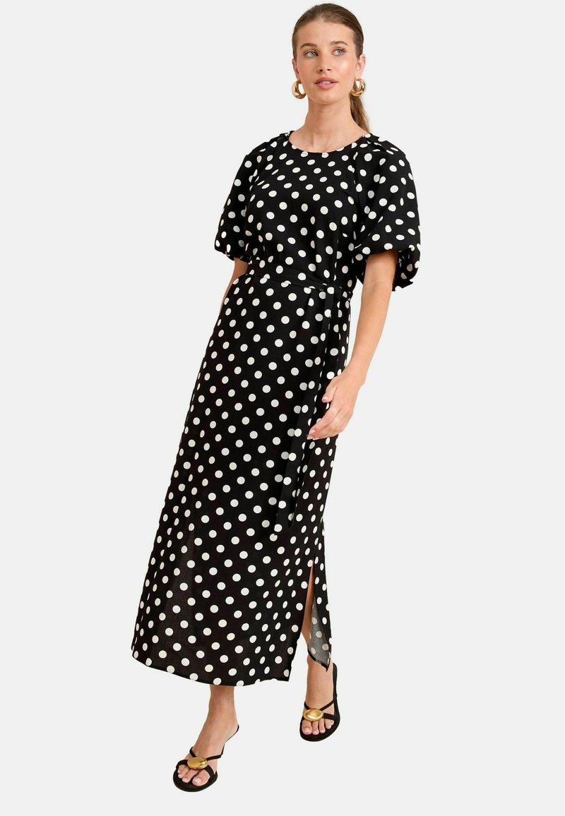 Rochie maxi neagră cu buline albe, mâneci scurte tip puff, cordon la talie și fanta laterală. Purta cu sandale negre cu accente aurii.