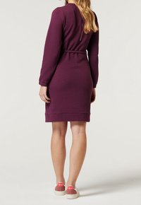 Vestido burdeos de manga larga con una silueta ajustada y cinturón. Tela texturizada, hasta la rodilla, combinado con zapatillas blancas y rojas.