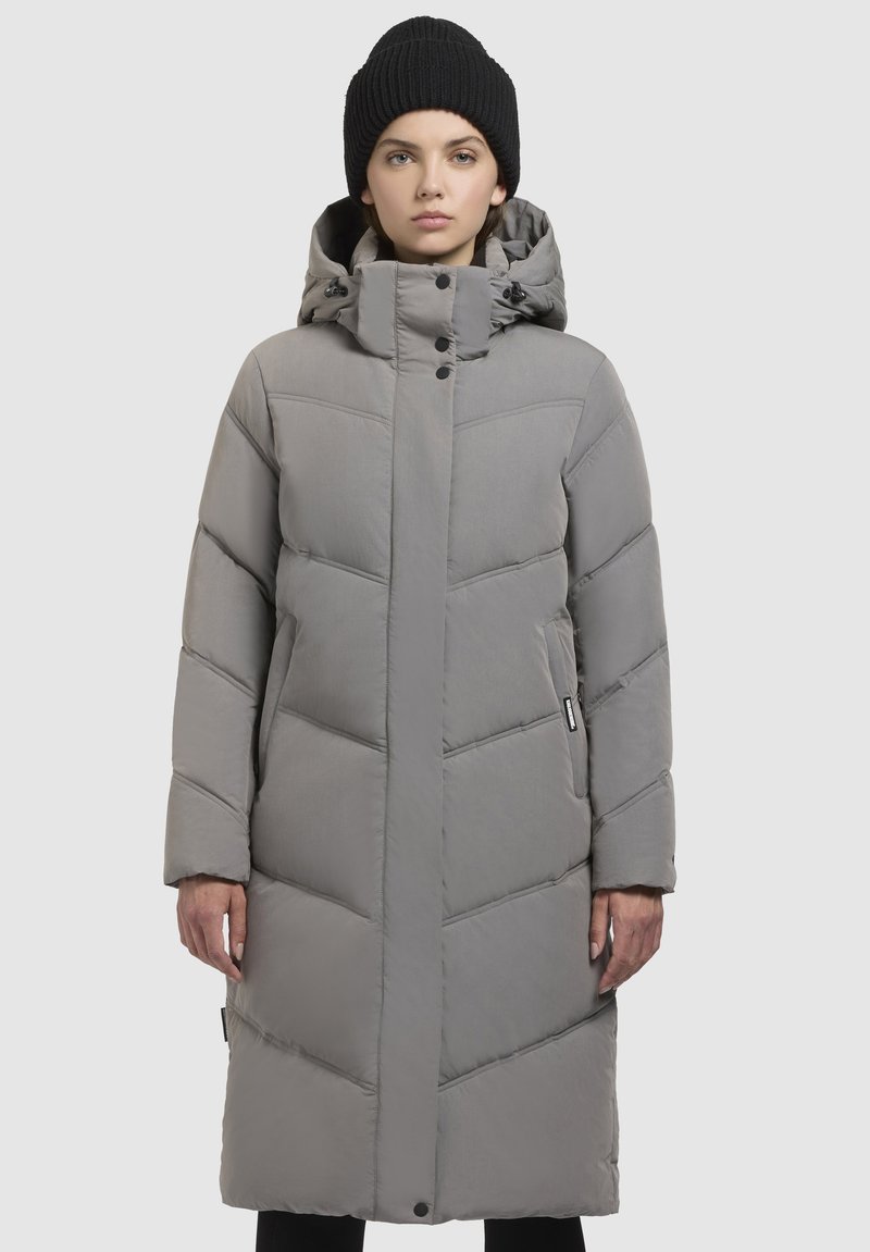 Abrigo largo acolchado gris con cuello alto, cierre de cremallera y botón a presión, con acolchado en forma de chevrón y bolsillos laterales. Combinado con un gorro negro.