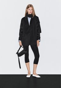 Donna che indossa un blazer nero, sciarpa e leggings, che tiene una borsa nera, in piedi contro un muro bianco e un pavimento nero.