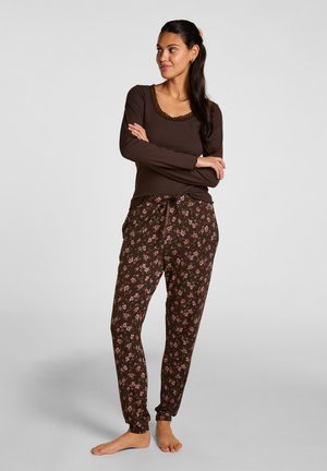 Donna con lunghi capelli scuri che indossa una maglia marrone a maniche lunghe e pantaloni da pigiama con fantasia floreale, in piedi a piedi nudi con le braccia incrociate.