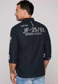 Camisa negra de manga larga con texto bordado en la parte posterior, con mangas enrollables con un acento a rayas y un diseño de corte holgado.