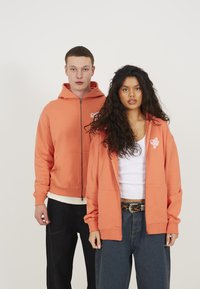 To modeller har på seg matchende korallfargede zip-up hettegensere laget av mykt materiale, med lommer foran og en logo på brystet. De kombinerer med mørke jeans.