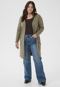 Kvinde iført en olivengrøn lang cardigan, mørkebrun top, blå lige ben jeans og sorte sko, stående med den ene hånd i lommen.