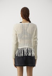 Dzianinowy kremowy sweter z otwartym splotem i frędzlami na dolnym brzegu, zestawiony z czarną spódnicą dżinsową. Posiada długie rękawy i luźny krój.