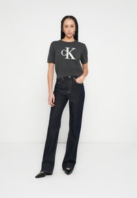 Tricou gri cu mâneci scurte și logo alb "CK", asortat cu blugi drepti, de culoare albastru închis. Modelul stă cu o mână în buzunar.