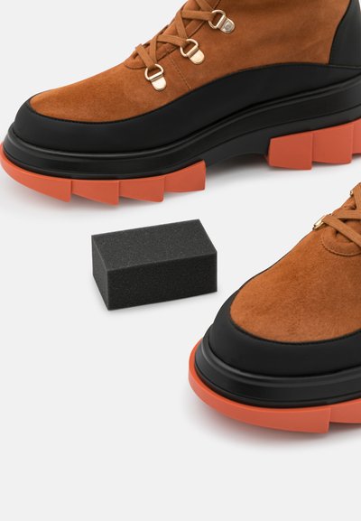 Brown suede ankle boots with black leather accents and chunky orange rubber soles; features gold eyelets and brown laces.