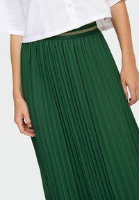 Gonna plissettata verde con finitura testurizzata, dotata di una cintura elastica con accenti dorati, abbinata a una maglietta corta bianca.