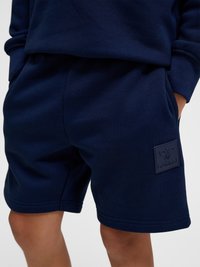 Personne portant un short bleu marine les mains dans les poches, avec un patch logo carré sur la cuisse gauche.