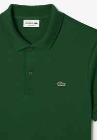 Groene polo met een gestructureerde stof, een kraag, drie knopen en een klein geborduurd krokodillogo op de linkerborst.