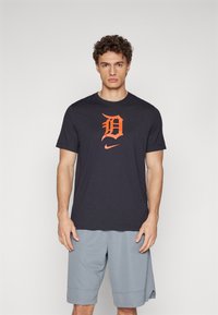 Tamsiai pilkas trumpomis rankovėmis marškinėliai su oranžiniu "D" logotipu ir Nike swoosh, pagaminti iš minkštos medžiagos, su įprastu kirpimu.