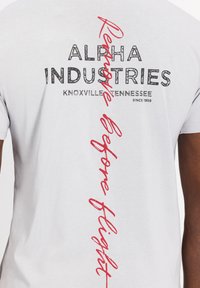 Siva bomba majica iz bombažnega materiala z drzno črno oznako "ALPHA INDUSTRIES", podrobnostjo "KNOXVILLE TENNESSEE" in rdečim besedilom "Remove Before Flight".