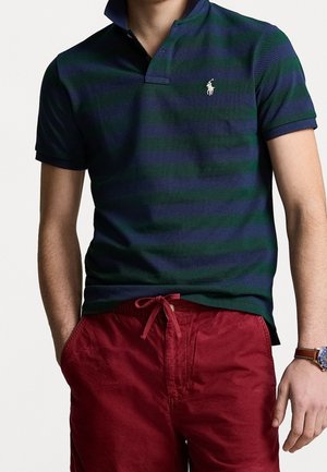 Man wearing a navy and dark green striped polo shirt with a white polo player logo, red drawstring shorts, and a watch on his left wrist.