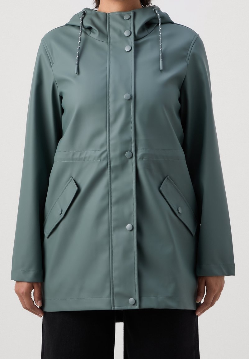 Femme portant un imperméable à capuche bleu sarcelle avec fermeture à boutons sur le devant et poches à rabat, debout devant un fond uni.